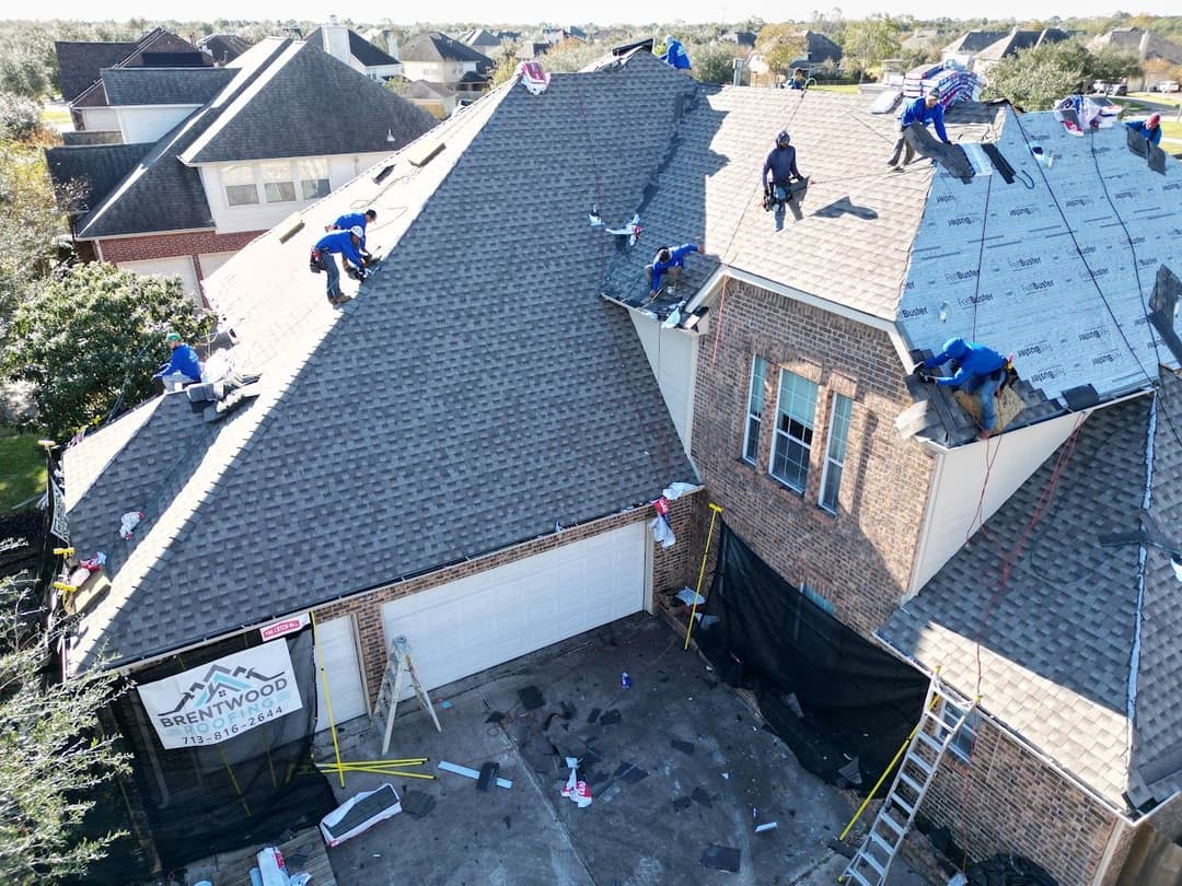 Roofers installing shingles on a residential home with a sign for Brentwood Roofing Services.