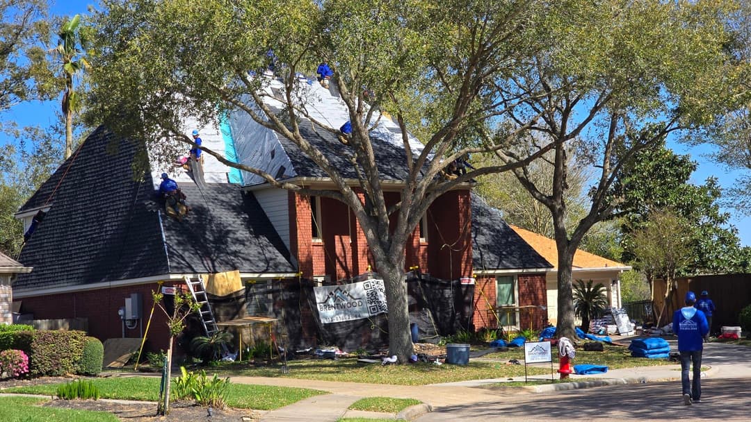 Roofing team working on a house with a black shingle roof, surrounded by trees and landscaping.