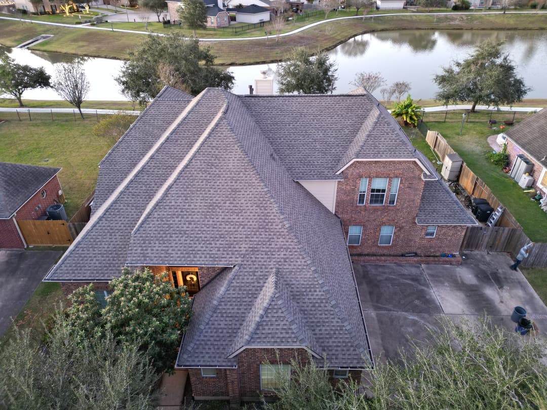 Aerial view of a brick home with a grey roof near a pond and landscaped yard.