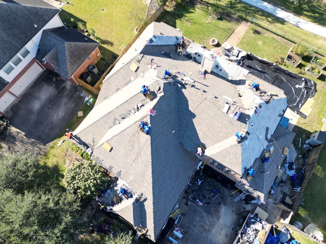 Aerial view of a house undergoing roofing repairs with workers on the roof and materials scattered.